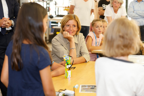 In der Mitte des Bildes sitzt Ministerpräsidentin Malu Dreyer an einem Tisch; sie blickt auf ein Mädchen, das mit dem Rücken zur Kamera steht. Weitere Kinder und eine Erwachsene sieht man im Hintergrund.