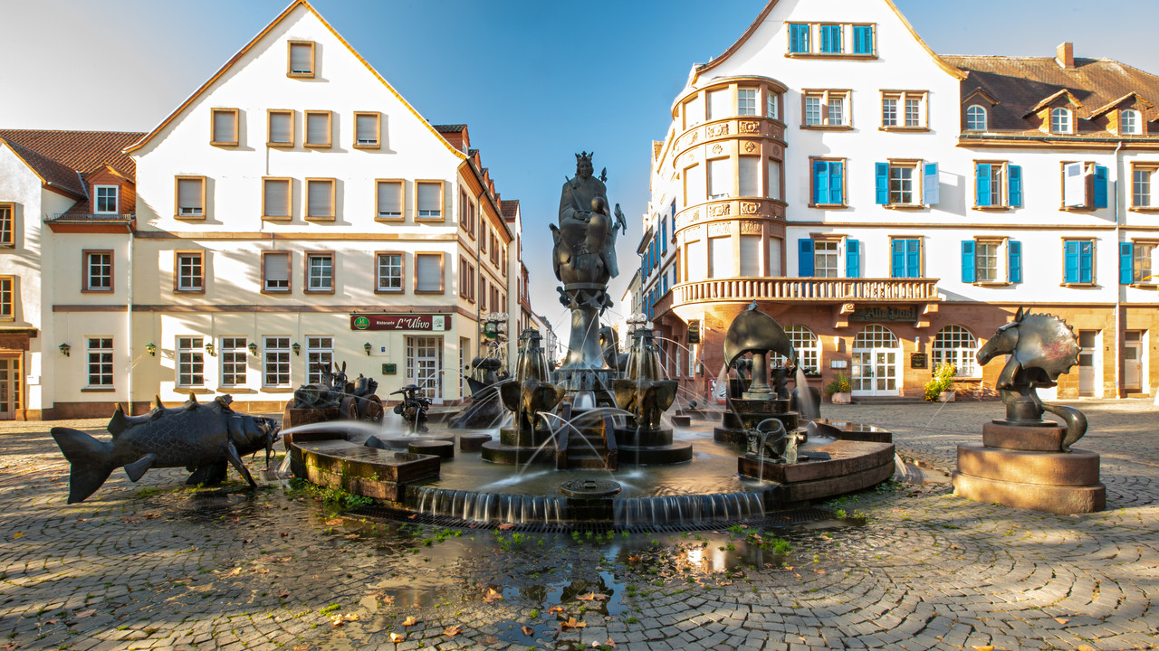 Kunstvoller Brunnen auf einem sonnigen Marktplatz, umgeben von historischen Häusern mit blauen Fensterläden.