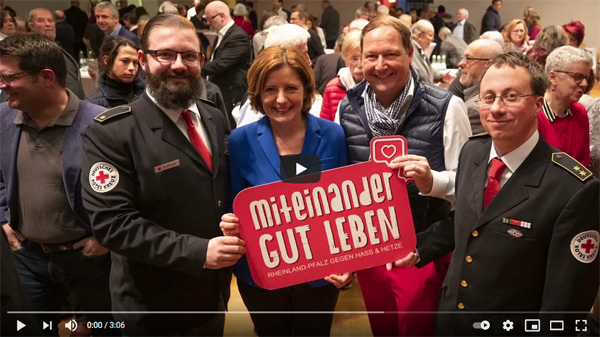Vier in die Kamera blickende Menschen, inkl. Ministerpräsidentin Malu Dreyer; sie halten ein Schild mit der Aufschrift „Miteinander gut leben“ Dahinter sind weitere Menschen zu sehen.